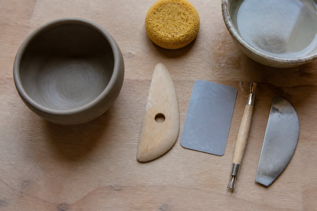Overhead view of a pottery studio table with a finished clay pot, and next to it a collection of repurposed tools like a wooden rib, a credit card for scraping, and a needle tool, with a sponge and water bowl nearby