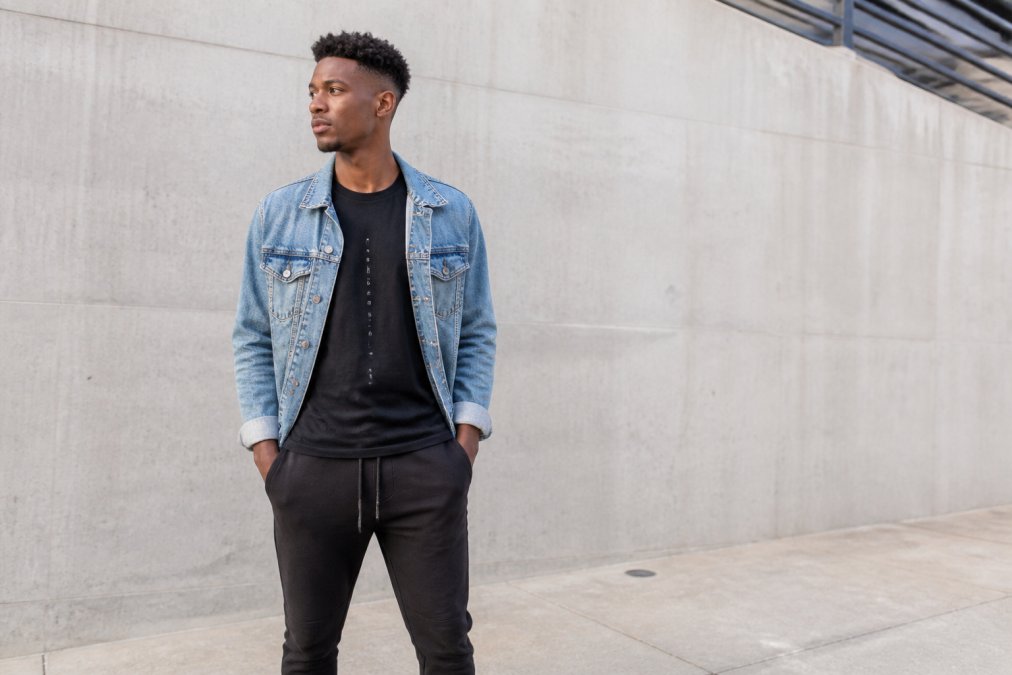 A young adult wearing a modern graphic tee with subtle faith-inspired lettering, styled with a denim jacket and joggers, standing against a clean urban wall in soft daylight