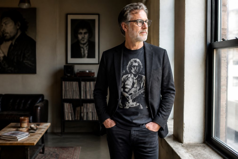 Man in his 50s wearing a vintage-inspired graphic tee with a charcoal blazer and dark jeans, standing in natural window lighting with confident posture