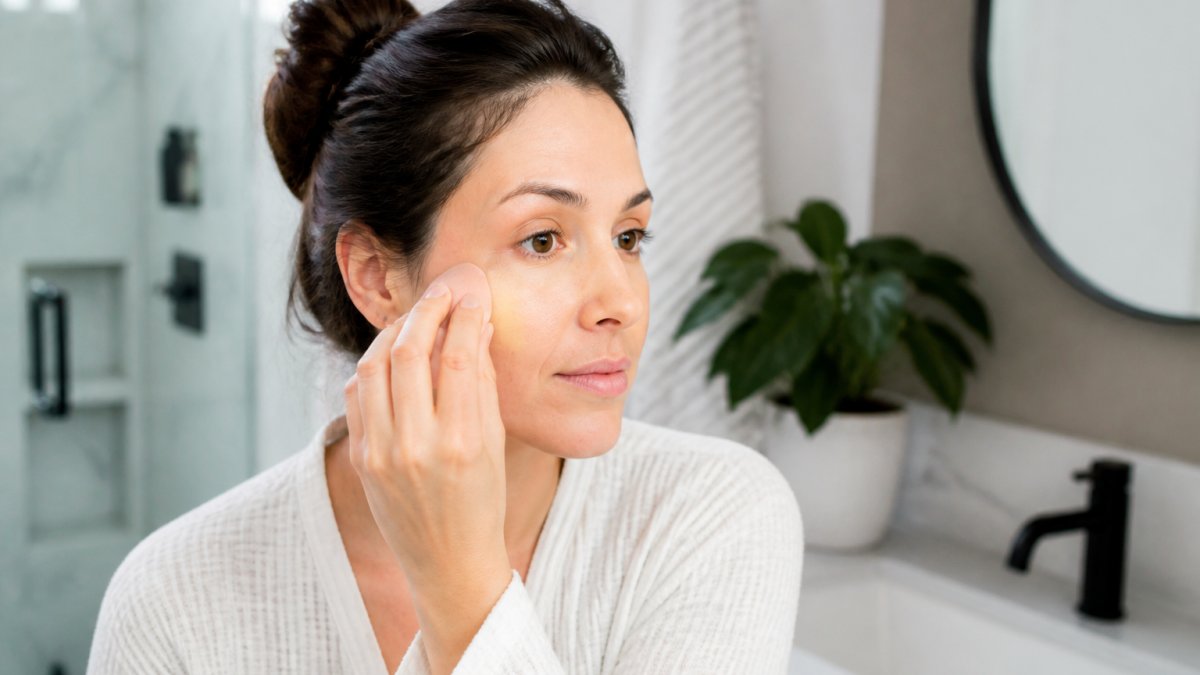 Woman with sensitive skin applying liquid foundation with a damp makeup sponge in a bright bathroom