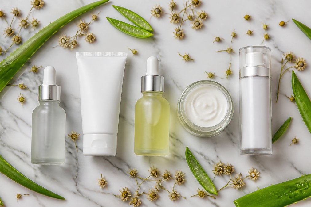 Flat lay of five clean skincare products arranged on a white marble surface, surrounded by fresh aloe vera leaves and small dried chamomile flowers, photographed in soft natural daylight from above