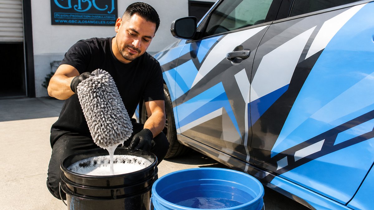 Professional detailer holding soft microfiber wash mitt over a bucket of soapy water next to a vehicle wrap, with cool rinse water bucket visible in natural outdoor lighting
