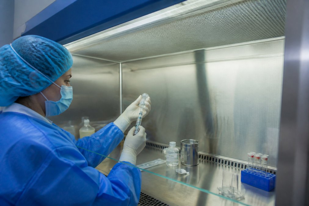 Pharmaceutical technician performing sterile compounding inside a Class II biosafety cabinet, hands working at the laminar airflow work surface with proper gowning, clear HEPA filter visible above