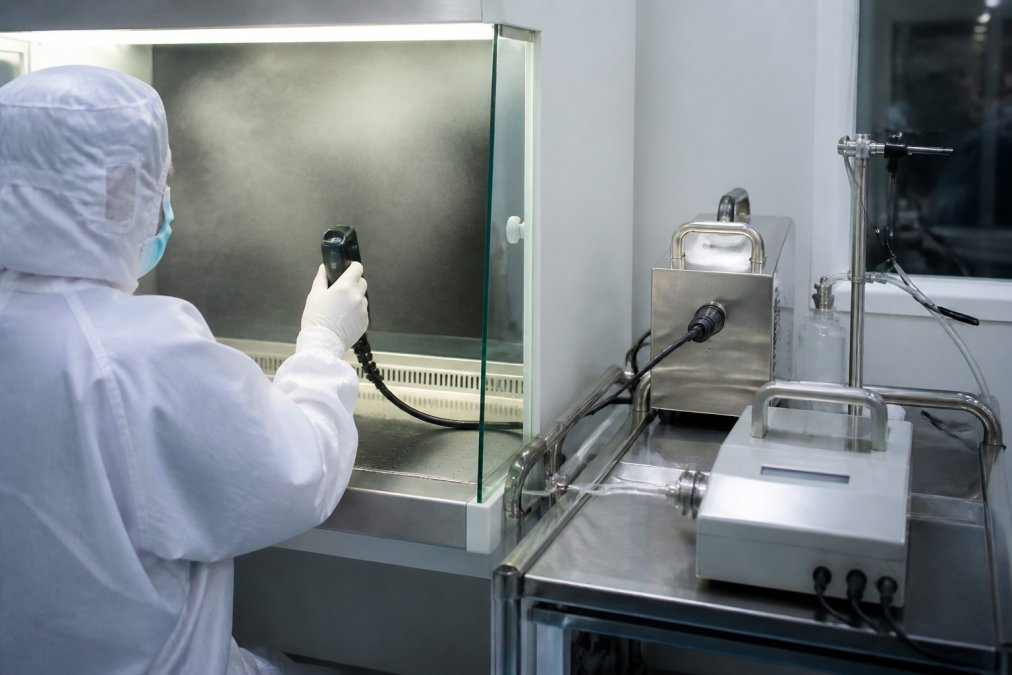 Cleanroom validation specialist using aerosol generator and photometer equipment to test HEPA filter integrity and laminar airflow patterns, holding scanning probe near biosafety cabinet work surface with visible aerosol cloud under controlled lighting