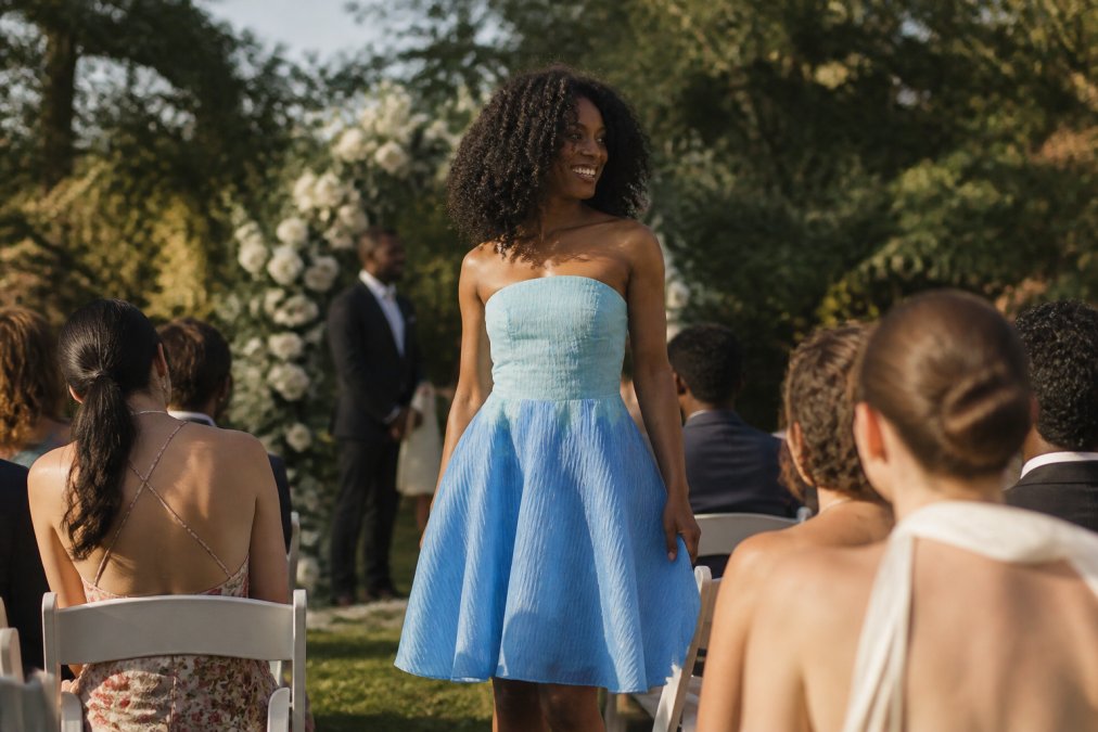 Woman wearing a vibrant blue ombré organza dress at an outdoor garden wedding ceremony, with soft natural light catching the luminous gradient effect as she stands among seated guests, showcasing how the dress photographs beautifully in daylight