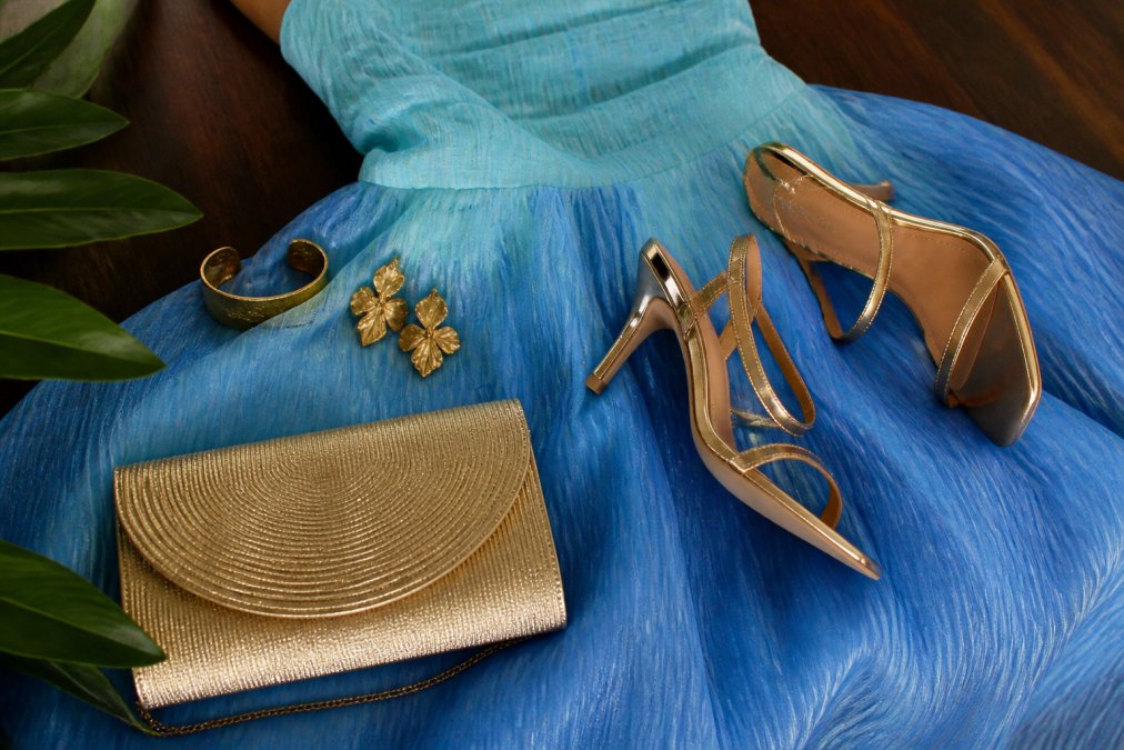 Close-up detail shot of gold jewelry, strappy heels, and a metallic clutch arranged on jewel-toned blue ombré fabric, showing how understated metallic accessories complement bold occasion wear without overwhelming it