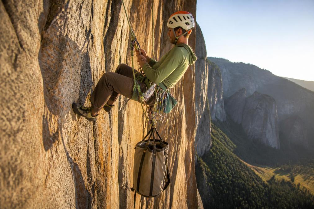 Climber hanging on a big wall route wearing a padded climbing harness loaded with gear above a suspended haul bag.