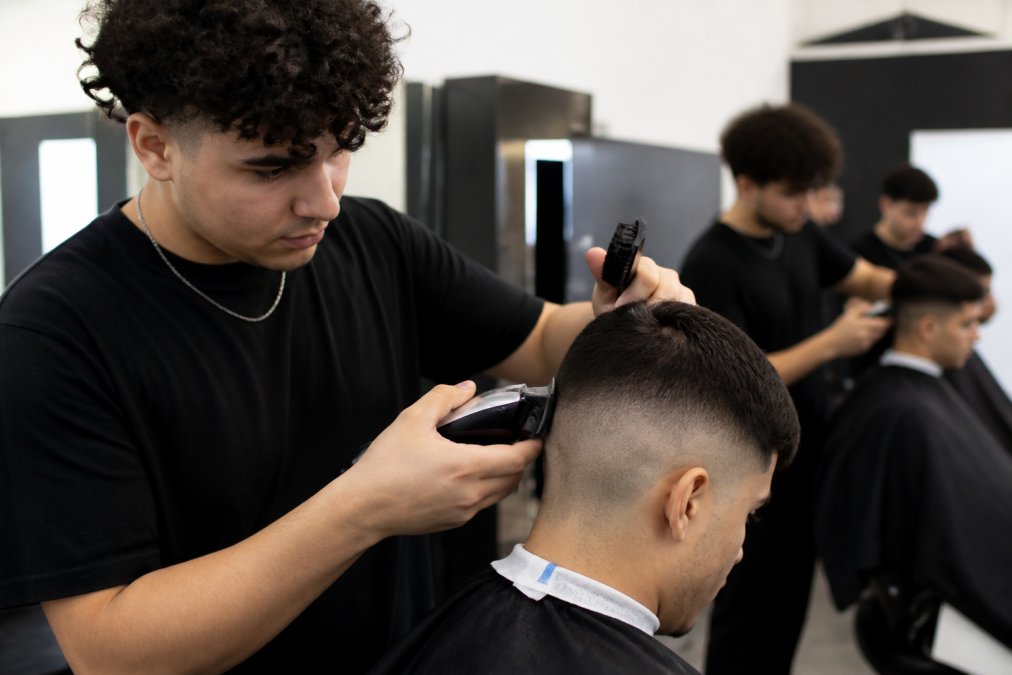 A barber student in a training environment holding professional clippers with proper grip and positioning over a client's head during a fade cut, with natural studio lighting