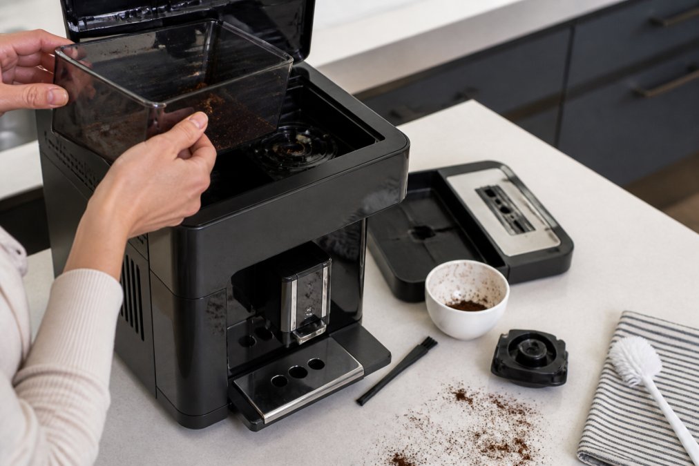 Close-up of hands removing and brushing out the bean hopper of a super-automatic espresso machine on a clean kitchen counter, with a small cleaning brush and scattered coffee grounds visible, bright overhead kitchen lighting