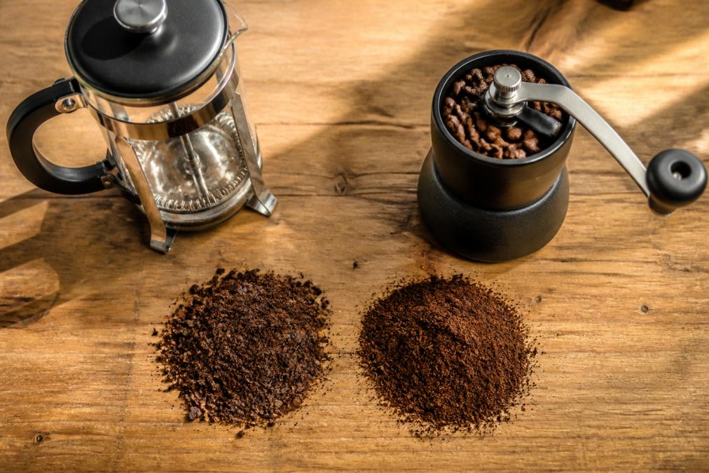 Overhead close-up of two piles of coffee grounds on a wooden counter, one coarse and one fine, beside a glass French press and a manual burr grinder in warm morning light