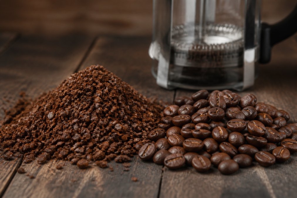 Close-up of coarse ground coffee beans next to whole beans on a dark wooden table, with a French press carafe blurred in the background