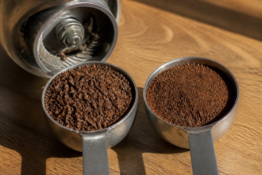 Close-up of coarse coffee grounds in a measuring scoop next to a burr grinder on a wooden counter with morning sunlight