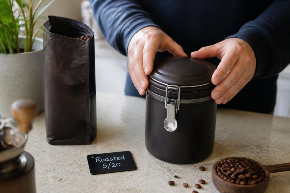 A person pressing the lid closed on a dark airtight ceramic coffee canister on a kitchen counter, with an open bag of whole bean coffee nearby and a small chalkboard label showing a roast date written in chalk