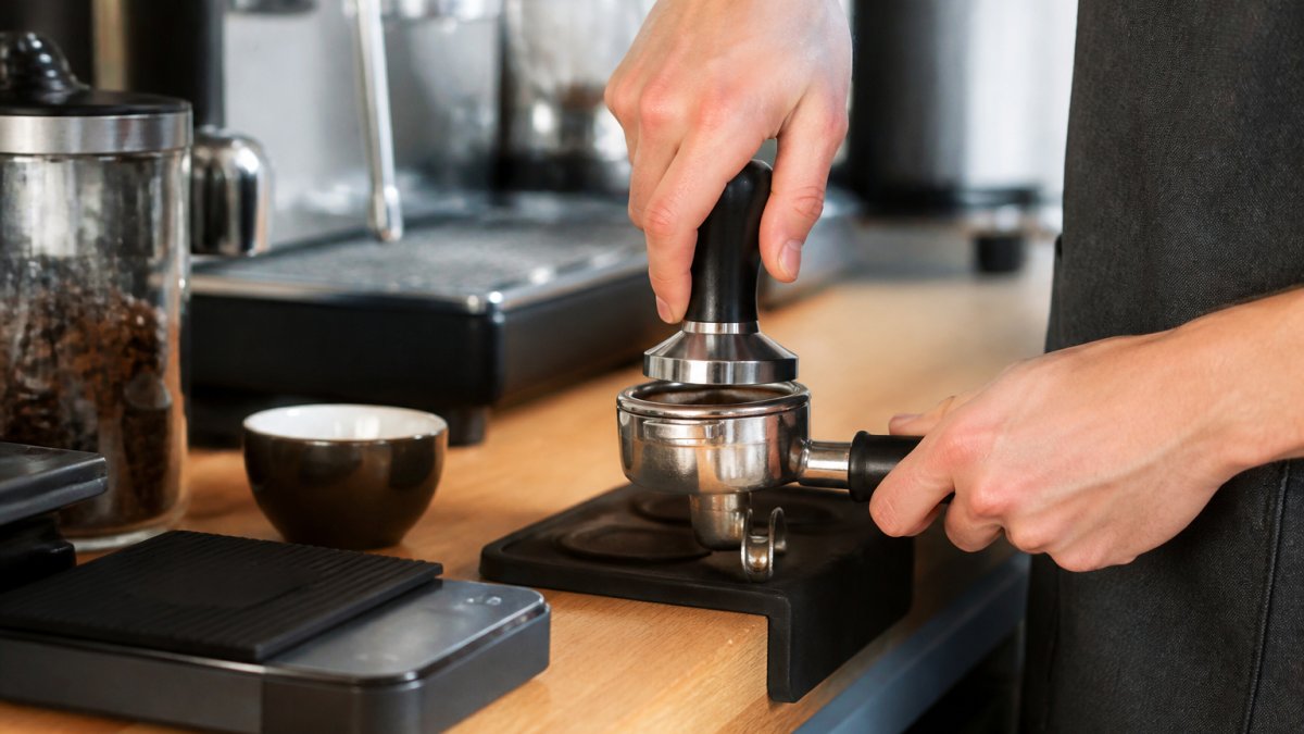 Close-up of a home barista's hands tamping freshly ground coffee into a portafilter on a kitchen counter, with a digital scale and espresso cup nearby