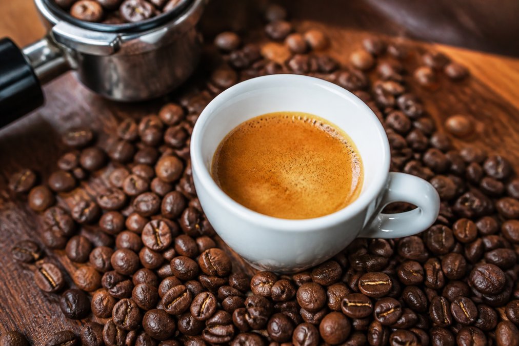 Close-up of a freshly pulled espresso shot in a small white ceramic cup, with a thick golden-brown crema on top, surrounded by whole coffee beans on a wooden surface
