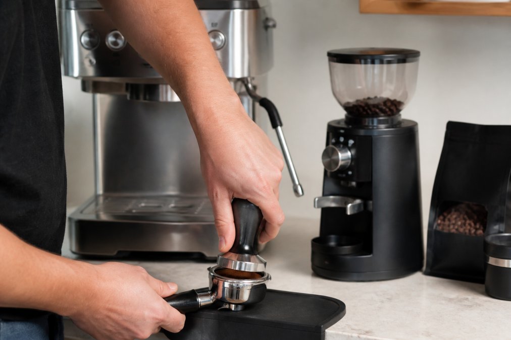 Hands tamping finely ground espresso into a portafilter on a home espresso machine, with a small grinder and bag of whole beans visible in the background on a kitchen counter