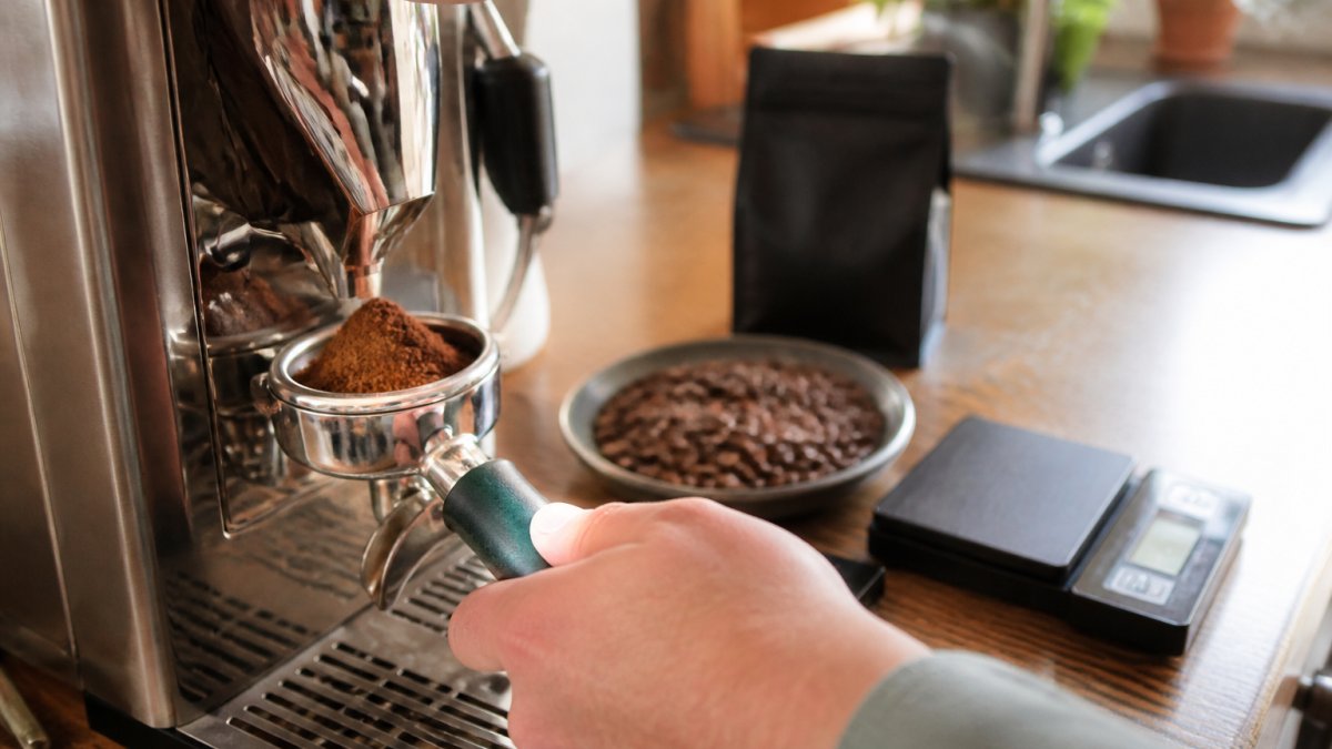 Close-up of a home espresso machine portafilter being filled with freshly ground coffee, with a bag of whole beans and digital scales on a kitchen counter