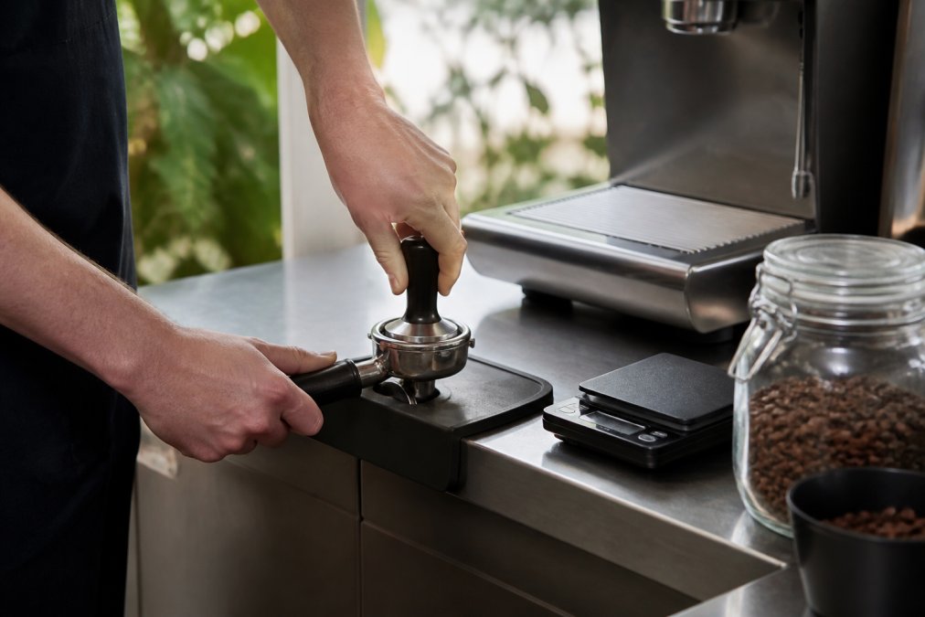 Home barista tamping the best coffee beans for home espresso into a portafilter on a scale in a bright kitchen.