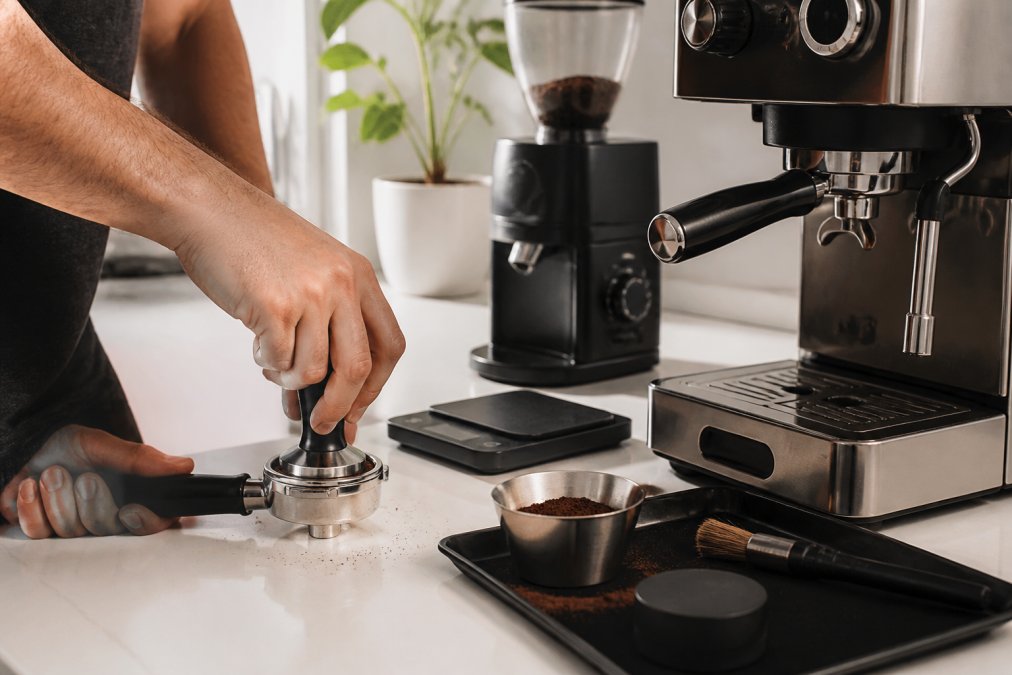 Hands tamping freshly ground espresso into a portafilter on a home espresso machine, small digital scale and burr grinder visible in the background on a clean white kitchen counter, warm natural light