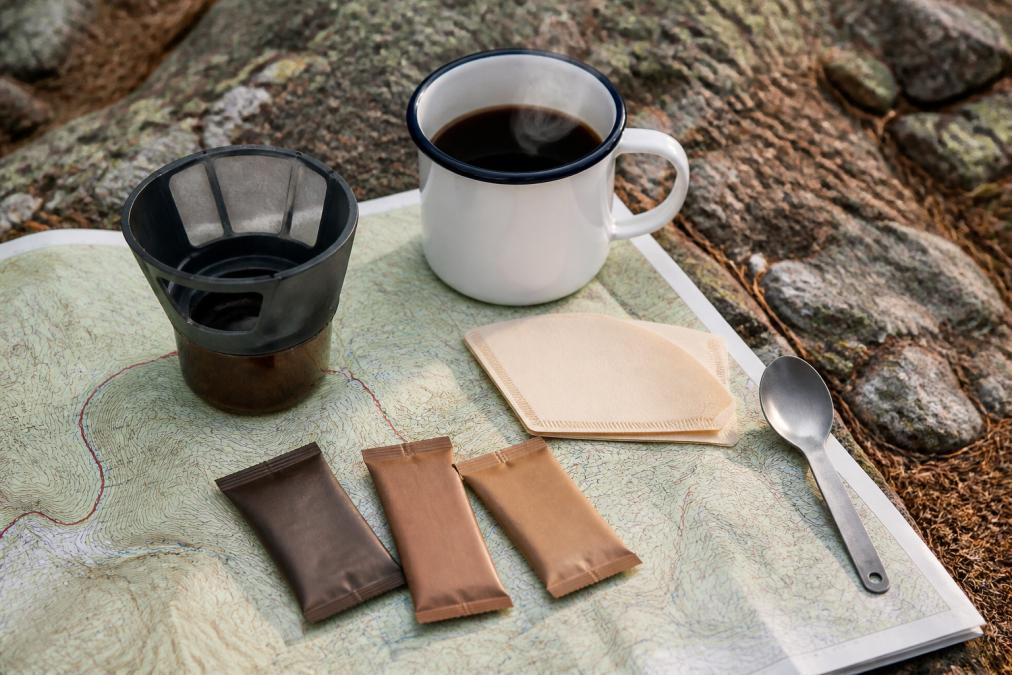 Flat lay of ultralight backpacking coffee gear arranged on topographic map: GSI Ultralight Java Drip, individual instant coffee packets, paper filters, metal camp spoon, and enamel mug with steam rising from rim