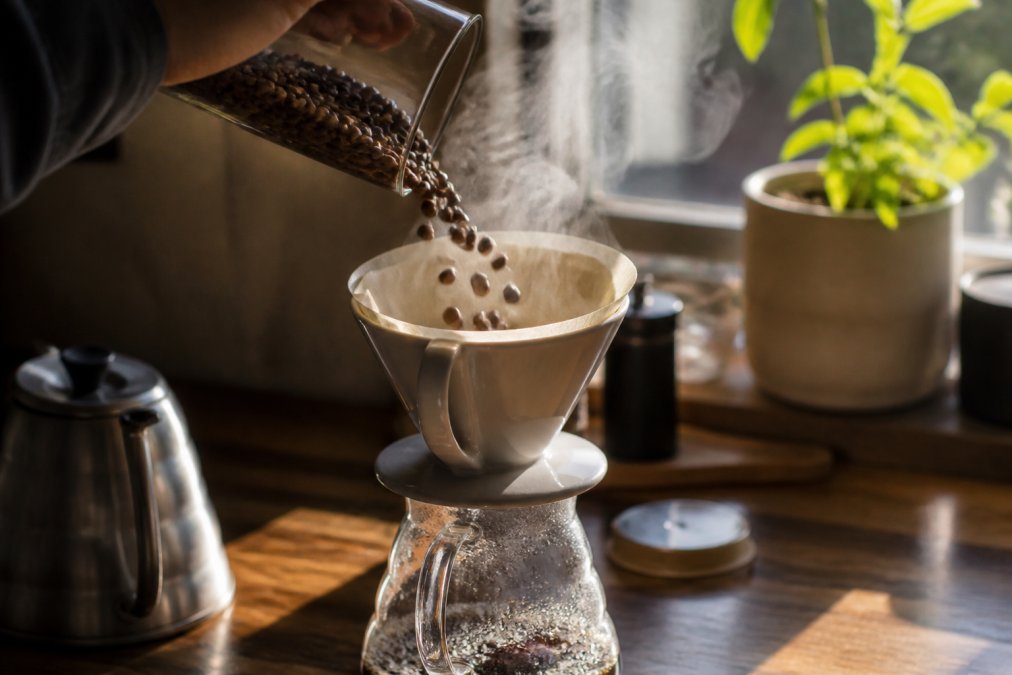 Close-up of whole coffee beans being poured into a ceramic pour-over cone with steam rising, morning sunlight streaming through a kitchen window