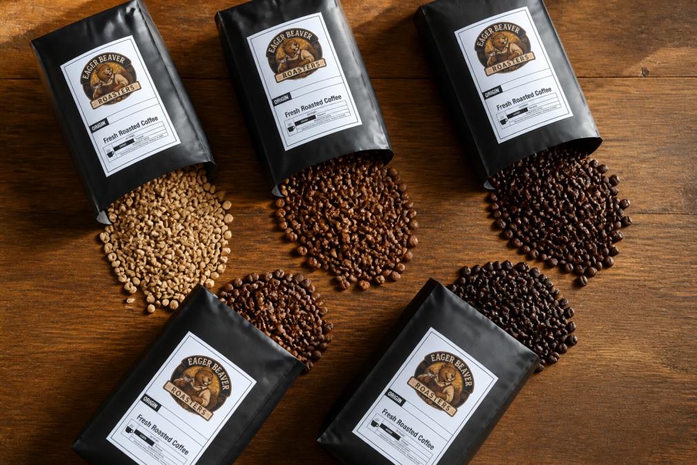 Overhead view of five different coffee bean bags arranged on a wooden table with fresh roasted beans spilling out onto the surface, showing variety of roast colors from light to dark brown, natural afternoon light highlighting the beans' texture and color differences