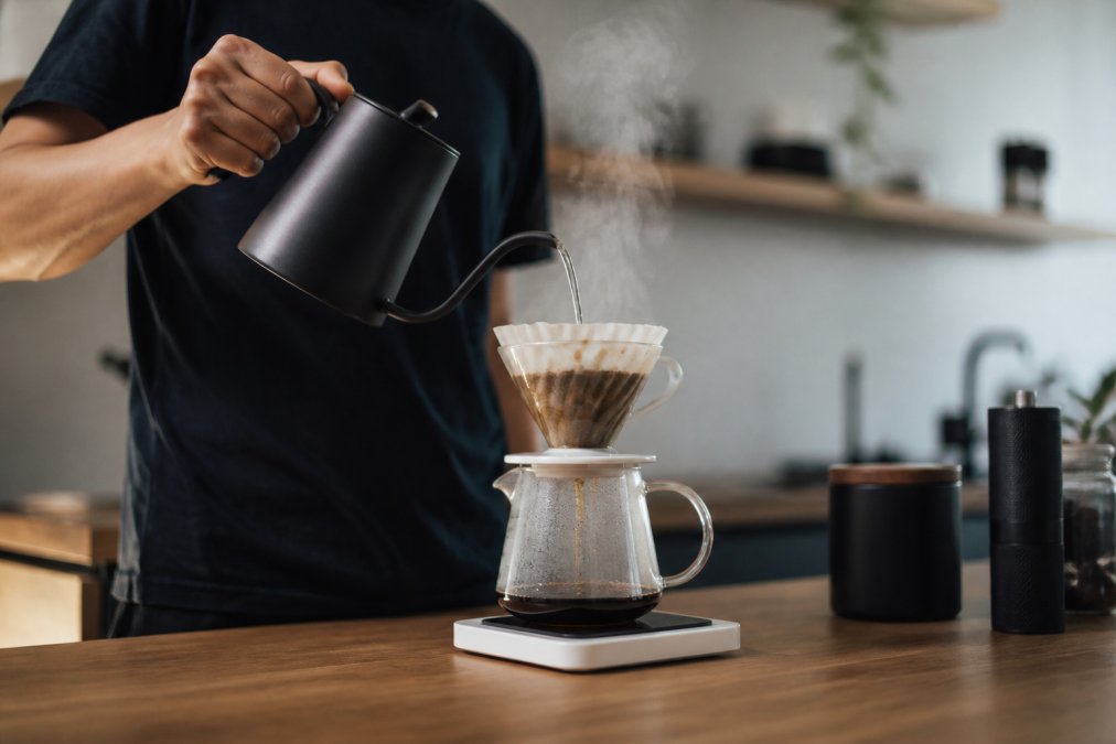 A person pouring hot water from a gooseneck kettle over a pour-over dripper filled with coffee grounds, steam rising, on a wooden kitchen counter with a digital scale nearby
