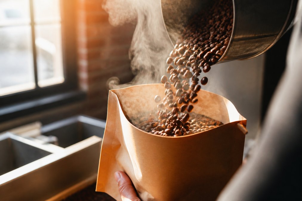 Close-up of freshly roasted coffee beans being poured into a kraft paper bag with morning sunlight streaming through a window, steam rising from the beans