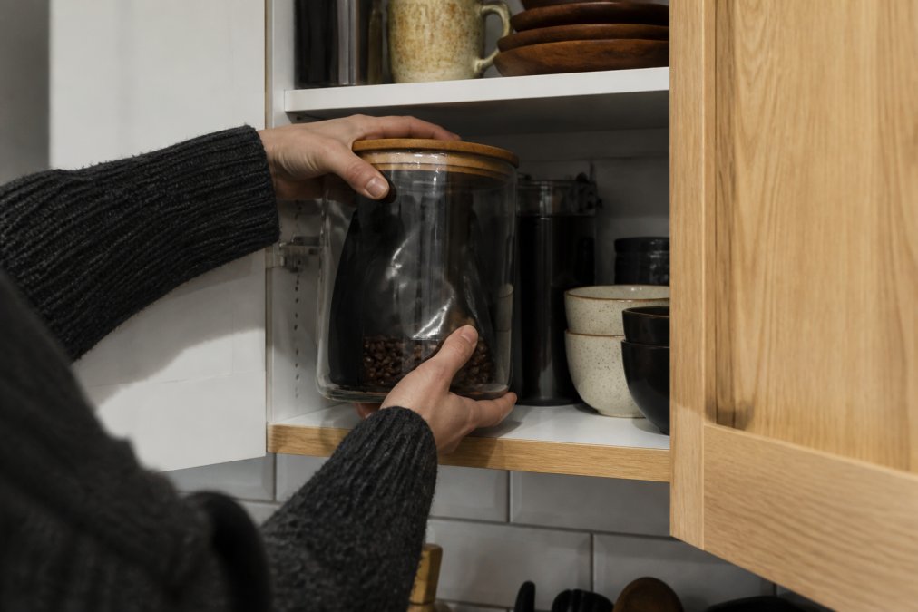 Hands placing opened coffee bag into an opaque airtight glass container in a kitchen cabinet, away from sunlight and heat sources, organized storage space visible