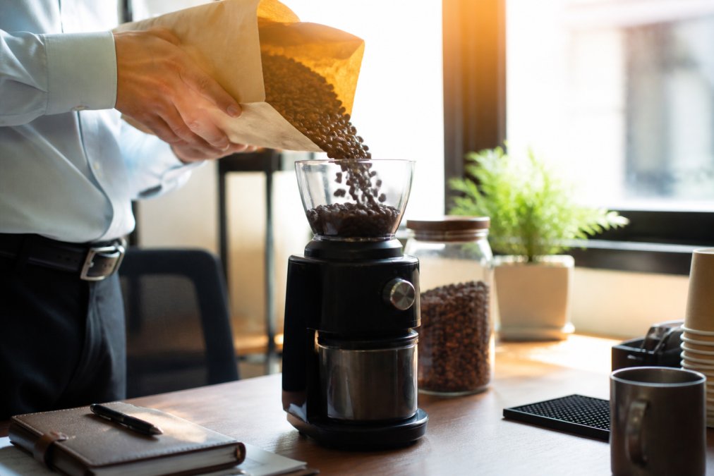 Hands pouring freshly roasted single-origin coffee beans from brown paper bag into electric grinder on clean desk, morning sunlight streaming through office window