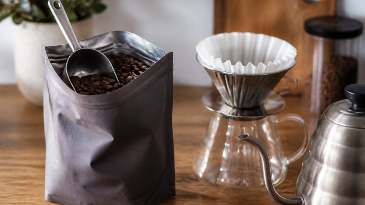 A close-up of a bag of coffee beans with a scoop, sitting on a wooden kitchen counter next to a pour-over setup