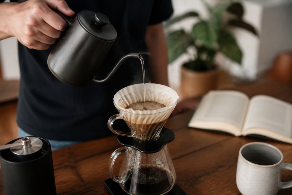 Close-up of hands pouring hot water over coffee grounds in a pour-over dripper, water streaming through the filter, with coffee equipment visible on a wooden counter and a book in soft focus in the background