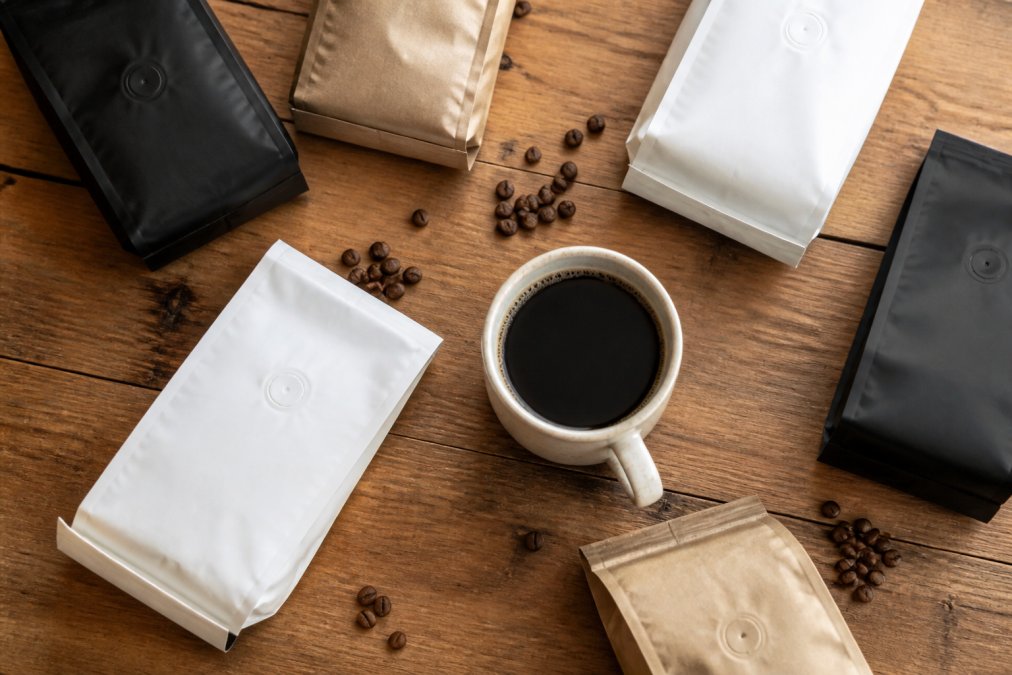 Overhead flat-lay of several whole-bean coffee bags arranged on a wooden table alongside a ceramic mug of black coffee, with a few scattered coffee beans on a warm-toned wooden surface in natural daylight