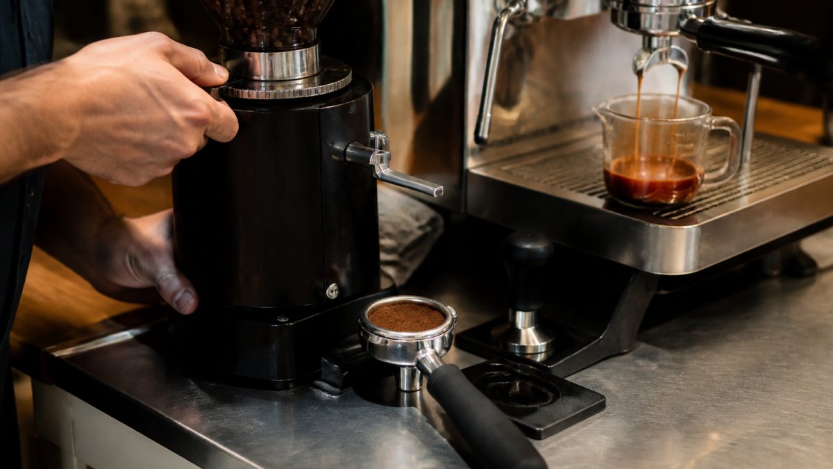 Close-up of barista's hands adjusting the burr grinder dial while a portafilter with espresso puck sits nearby, espresso shots flowing into a glass measuring cup on a stainless steel counter