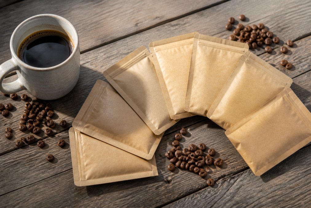 Flat lay of six small 2oz kraft paper coffee sample bags from Crystal Coast Coffee Co. arranged in a fan pattern on a weathered wooden table, with a white ceramic mug and scattered whole coffee beans, warm natural morning light