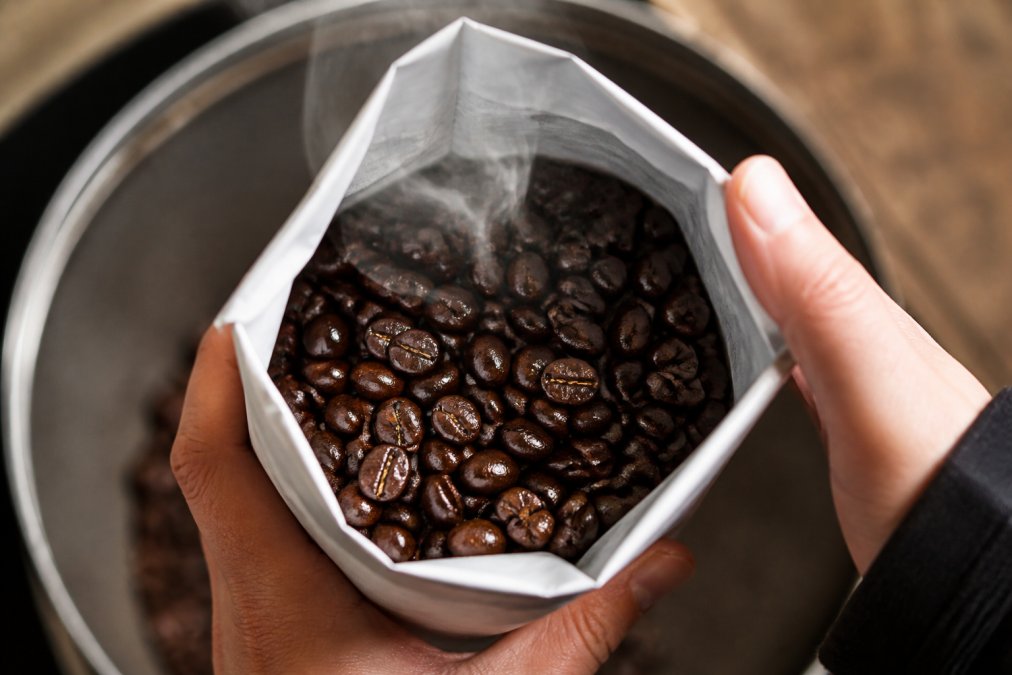 Close-up of freshly roasted coffee beans in a white paper bag with visible steam rising, hands holding the bag open to reveal the glossy dark beans with fresh roast sheen