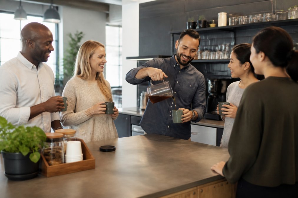 A diverse group of office employees standing around a modern breakroom counter, one person pouring coffee from a glass carafe into a mug while others chat and hold their own cups