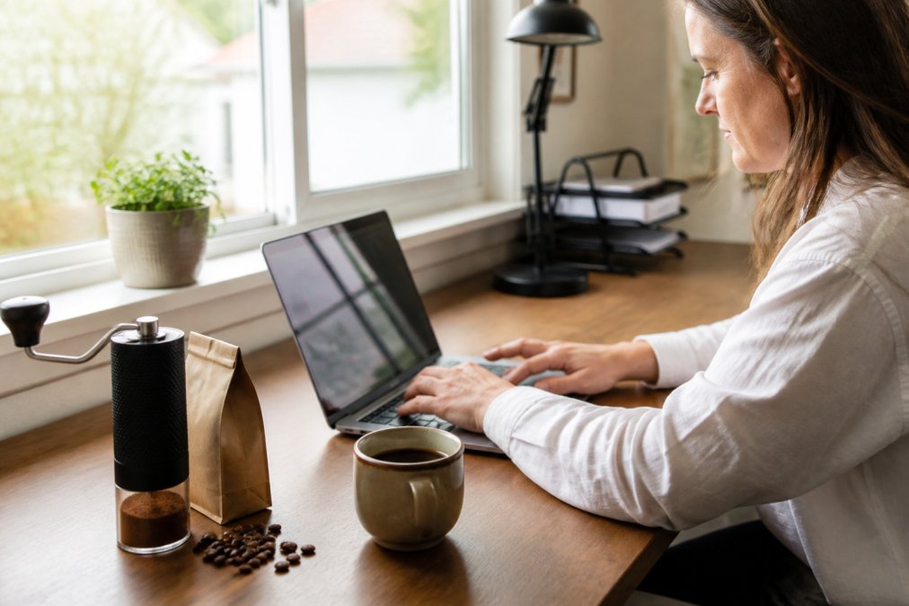 Remote worker grinding fresh beans for a coffee subscription at a bright home office desk with a laptop and mug.