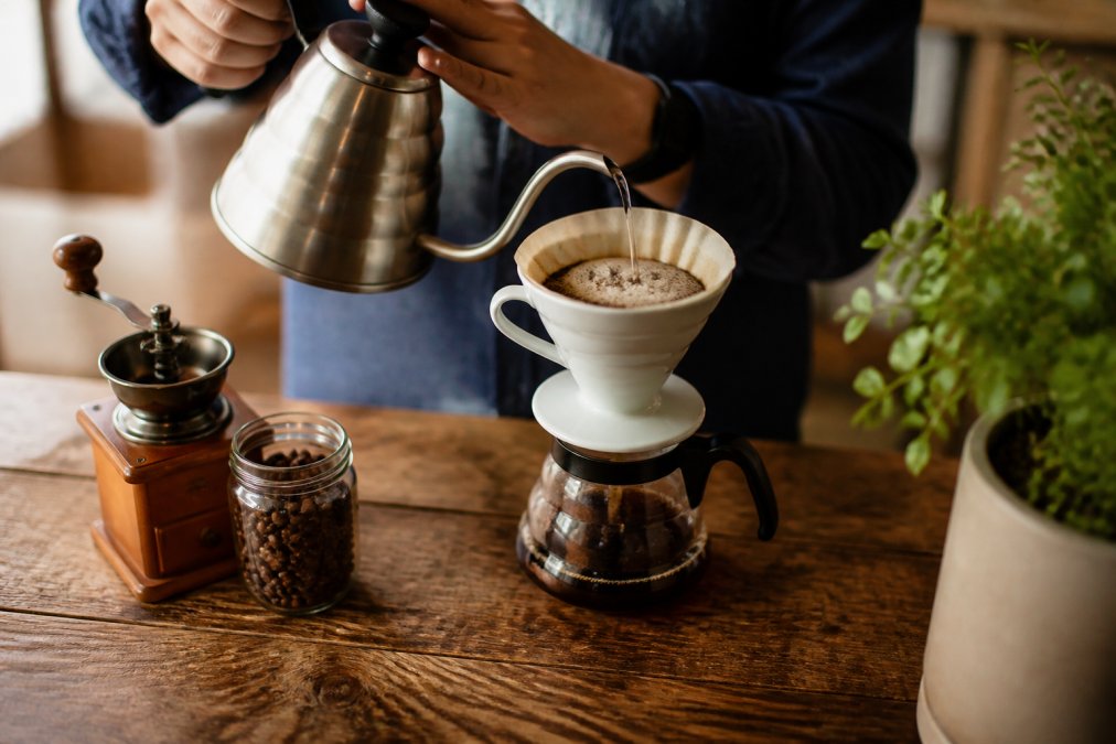 Close-up of hands pouring hot water from a gooseneck kettle over a pour-over coffee dripper on a wooden desk, with whole bean coffee and a small burr grinder visible in soft natural light nearby