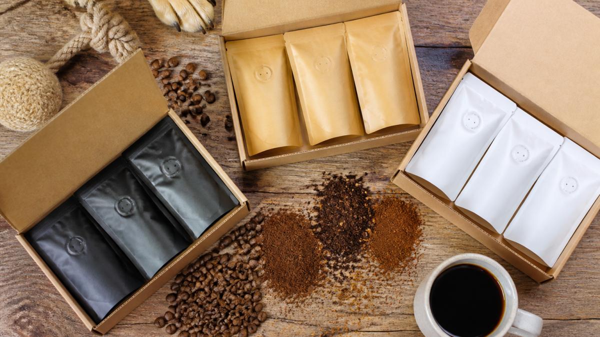 Overhead flat-lay of three open sample pack boxes displaying colorful coffee bags with whole beans and grounds spilled artfully across a wooden table, with a ceramic coffee cup and dog toy nearby in warm natural light