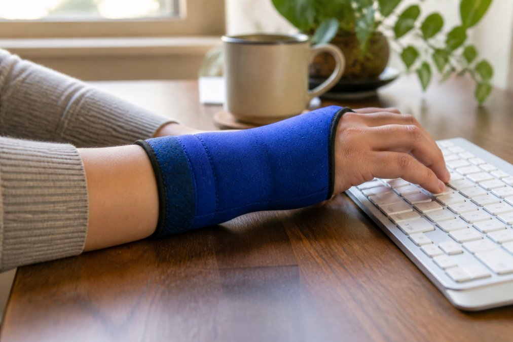 Close-up of a person's wrist wearing a fitted blue gel compression sleeve, resting on a desk beside a keyboard, soft natural light coming from a nearby window