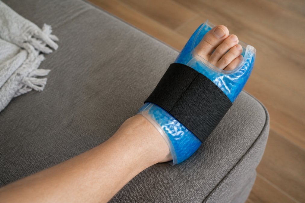 Person's foot wrapped with a flexible gel ice pack, showing proper application around the toe and forefoot area with visible compression, natural lighting from above