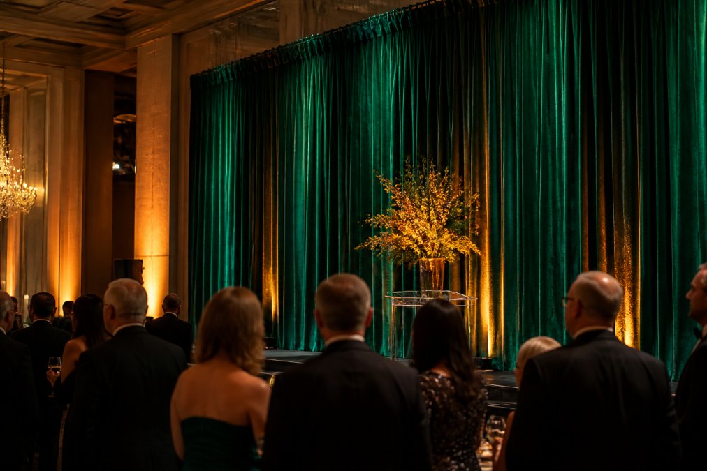 Elegant event backdrop featuring deep emerald velvet drape with warm amber stage lighting creating dramatic shadows and reflections, well-dressed attendees viewing the professional setup at a gala
