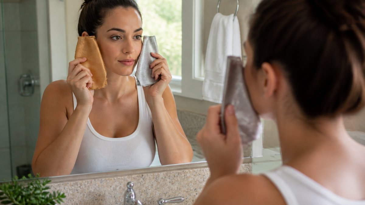 A woman in a bright bathroom holding two fabric swatches, one warm gold and one cool silver, next to her face while checking her reflection in a mirror