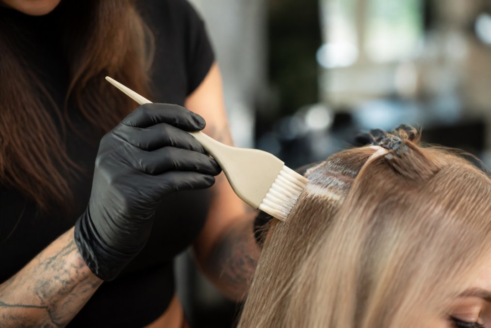 Professional hair colorist holding ESTL+CO feathered brush while applying precision color to client's hair, showing proper grip and controlled technique with natural salon lighting