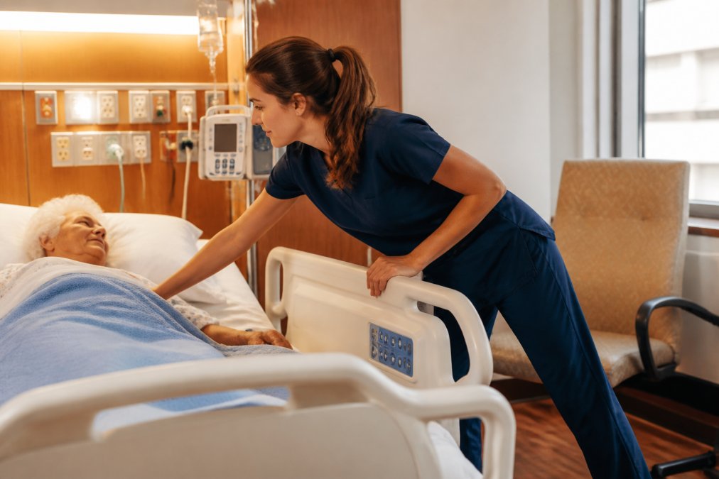 Nurse in comfortable scrubs bending and reaching across hospital patient bed during active clinical shift, demonstrating range of motion and fabric stretch in bright clinical lighting