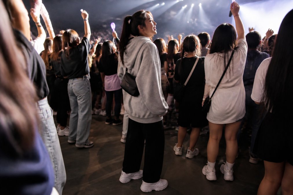 K-pop fan wearing comfortable athletic sneakers and oversized streetwear hoodie standing in crowded concert pit during active performance, surrounded by other fans with hands raised, stage lights illuminating the crowd from above