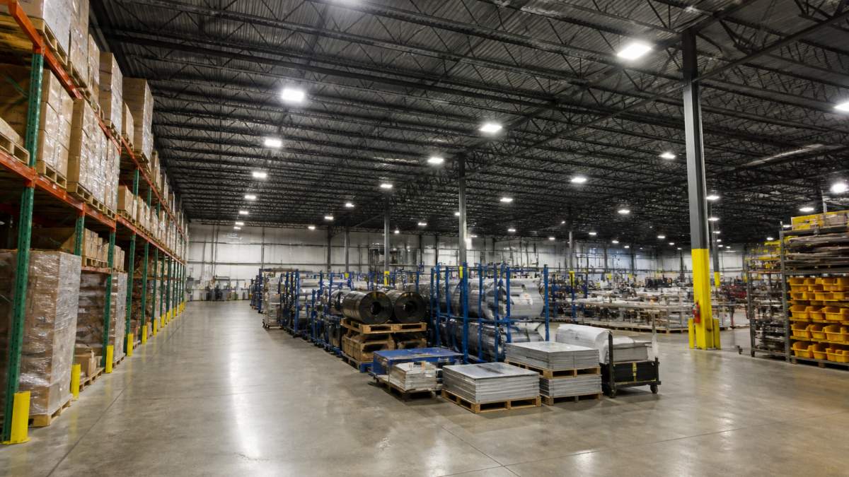 Wide-angle view of a modern warehouse interior with bright LED high-bay fixtures illuminating aisles of stored goods and equipment, metal shelving units, and industrial floor