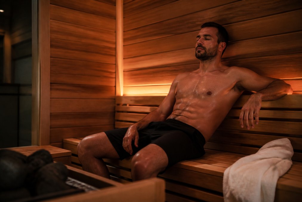 Member relaxing in modern commercial sauna with warm ambient lighting, wooden interior, towel draped nearby, showing premium wellness experience in clean professional environment