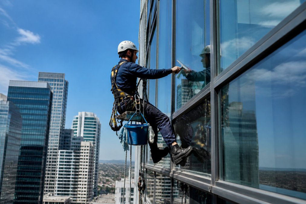 Professional window cleaning technician in full safety harness and fall protection equipment working on exterior glass panels of high-rise office building, suspended by rope access system against blue sky